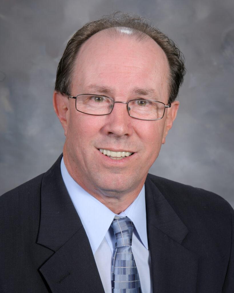 A middle-aged man with glasses, wearing a suit and tie, poses for a professional portrait against a gray backdrop.